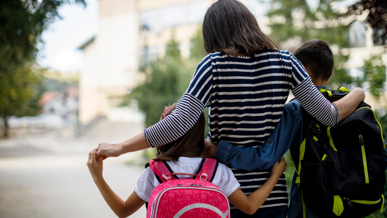 A parent is walking with two children, one on each side, down a pathway. The parent is wearing a striped shirt and has their arms around both children. The child on the left is carrying a pink backpack, and the child on the right is carrying a black and yellow backpack. The background shows a blurred view of buildings and trees, suggesting they are on their way to school.