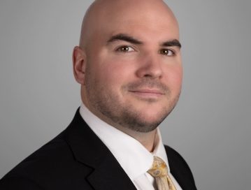 Patrick Apple Criminal Law Attorney Greensboro, NC; A professional headshot of Patrick Apple, a bald man with a trimmed beard. He is dressed in a dark suit, white shirt, and a gold patterned tie, looking confidently at the camera. The background is a neutral grey, providing a formal and polished appearance.