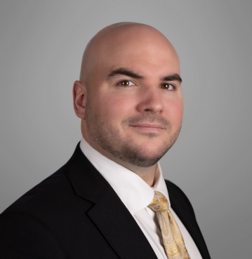 Patrick Apple Criminal Law Attorney Greensboro, NC; A professional headshot of Patrick Apple, a bald man with a trimmed beard. He is dressed in a dark suit, white shirt, and a gold patterned tie, looking confidently at the camera. The background is a neutral grey, providing a formal and polished appearance.