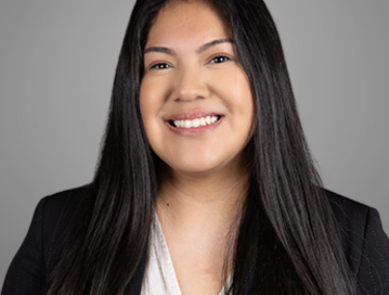 Diana Aguilar; A professional headshot of a young woman with long, straight black hair, smiling at the camera. She is wearing a black blazer over a white blouse. The background is a plain, light grey color, giving the photo a polished and corporate look.