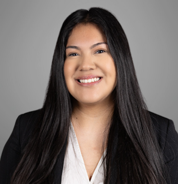 Diana Aguilar; A professional headshot of a young woman with long, straight black hair, smiling at the camera. She is wearing a black blazer over a white blouse. The background is a plain, light grey color, giving the photo a polished and corporate look.