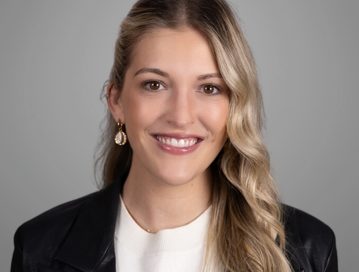 Molly Brazil Family Law Attorney Winston-Salem, NC; A professional headshot of a young woman with long, wavy blonde hair, styled with a side part. She is wearing a white top, a black blazer, and gold earrings. She is smiling warmly at the camera. The background is a neutral grey, adding to the formal and polished appearance of the photo.