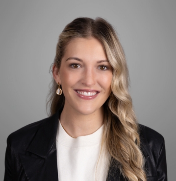 Molly Brazil Family Law Attorney Winston-Salem, NC; A professional headshot of a young woman with long, wavy blonde hair, styled with a side part. She is wearing a white top, a black blazer, and gold earrings. She is smiling warmly at the camera. The background is a neutral grey, adding to the formal and polished appearance of the photo.